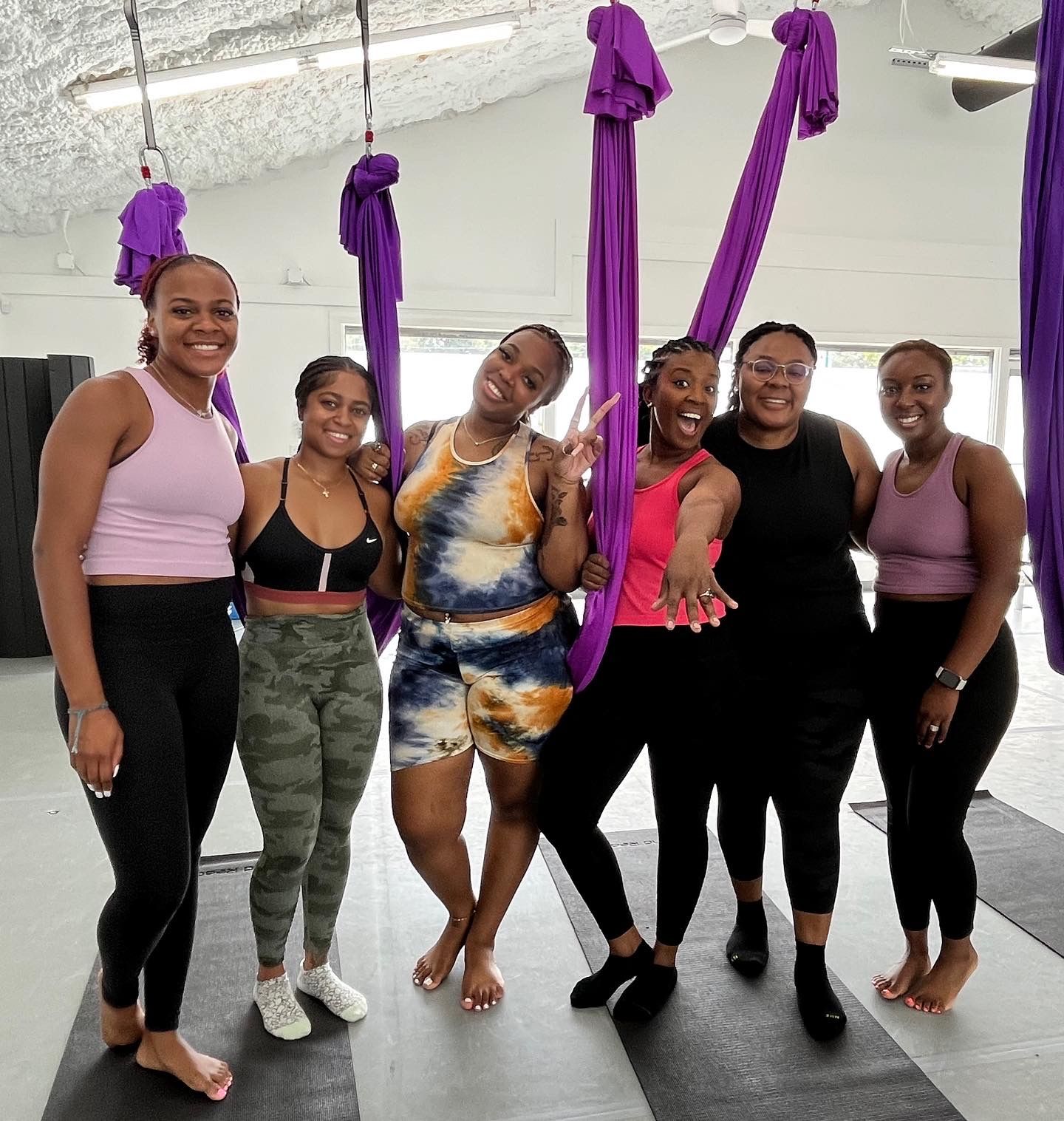 Six women in colorful activewear smiling and posing in an indoor aerial yoga studio with purple silk hammocks and black mats