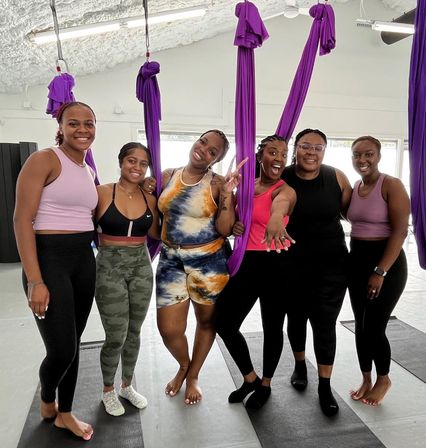 Six women in colorful activewear smiling and posing in an indoor aerial yoga studio with purple silk hammocks and black mats