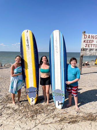 Three smiling beachgoers standing on a sandy shoreline holding long surfboards (yellow-striped and blue-striped) with the ocean and clear blue sky behind them and a weathered 'Danger Keep Off Jetty' sign visible