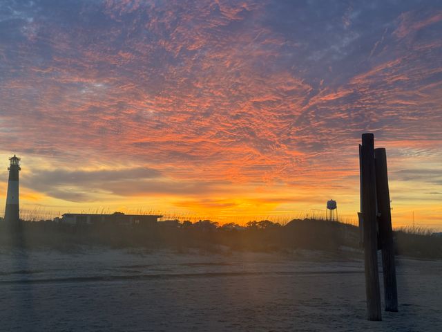 Stunning orange-pink sunrise over a sandy coastal beach with rippled clouds, a silhouetted lighthouse at left, wooden pilings at right, and grassy dunes with a distant water tower.