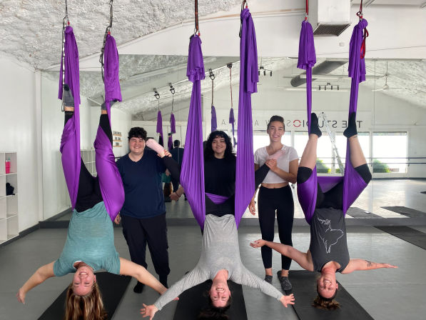 Group aerial yoga class in a bright studio with purple silks — three participants hanging upside down in hammocks while three others stand smiling by a mirrored wall