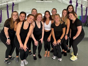 Smiling group of women in workout clothes posing in a mirrored aerial yoga studio with purple hammocks hanging from the ceiling.