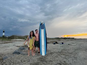 Two smiling beachgoers holding a blue-and-white surfboard on a sandy beach at sunset with a distant lighthouse, coastal dunes, and dramatic cloudy sky.