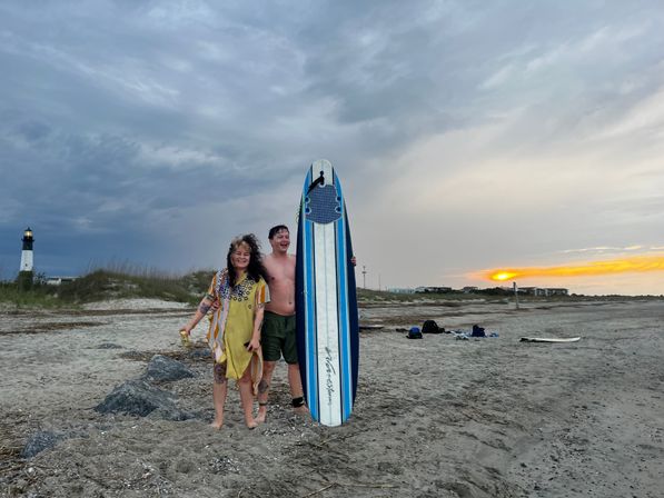 Two smiling beachgoers holding a blue-and-white surfboard on a sandy beach at sunset with a distant lighthouse, coastal dunes, and dramatic cloudy sky.