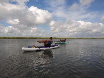 Two people balancing on paddleboards doing a playful core workout on calm estuary water with marsh grasses and a partly cloudy sky.