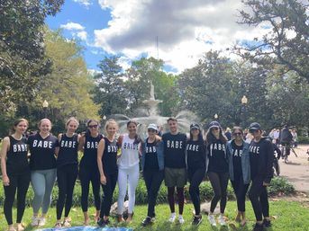 Bachelorette group wearing matching 'BABE' shirts with bride in white posing in front of a sunny park fountain