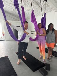 Three women smiling in an aerial yoga class holding purple silk hammocks above black mats and foam blocks in a bright fitness studio
