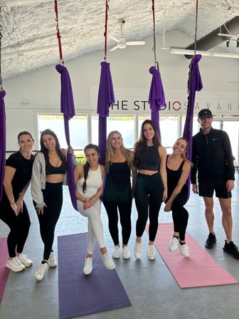 Seven smiling adults posing in a bright aerial yoga studio with purple silk hammocks suspended from the ceiling and colorful yoga mats on the floor, ready for a fitness class.