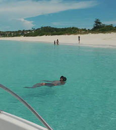 Person floating on their back in clear turquoise water near a white-sand beach with three people wading on shore, viewed from a boat railing under a bright blue sky