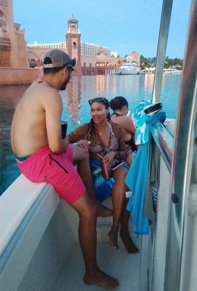 Three friends chatting on a boat at twilight in a calm marina, colorful towels on the rail, a smiling woman holding a drink, pink resort-style waterfront and yachts reflecting in the water.