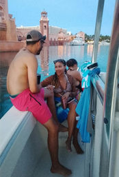 Three friends chatting on a boat at twilight in a calm marina, colorful towels on the rail, a smiling woman holding a drink, pink resort-style waterfront and yachts reflecting in the water.