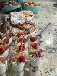 Brunch spread on a marble countertop: mini yogurt parfaits layered with granola, strawberries and raspberries in shot glasses with tiny spoons, beside powdered-sugar French toast and bowls of mixed fruit.
