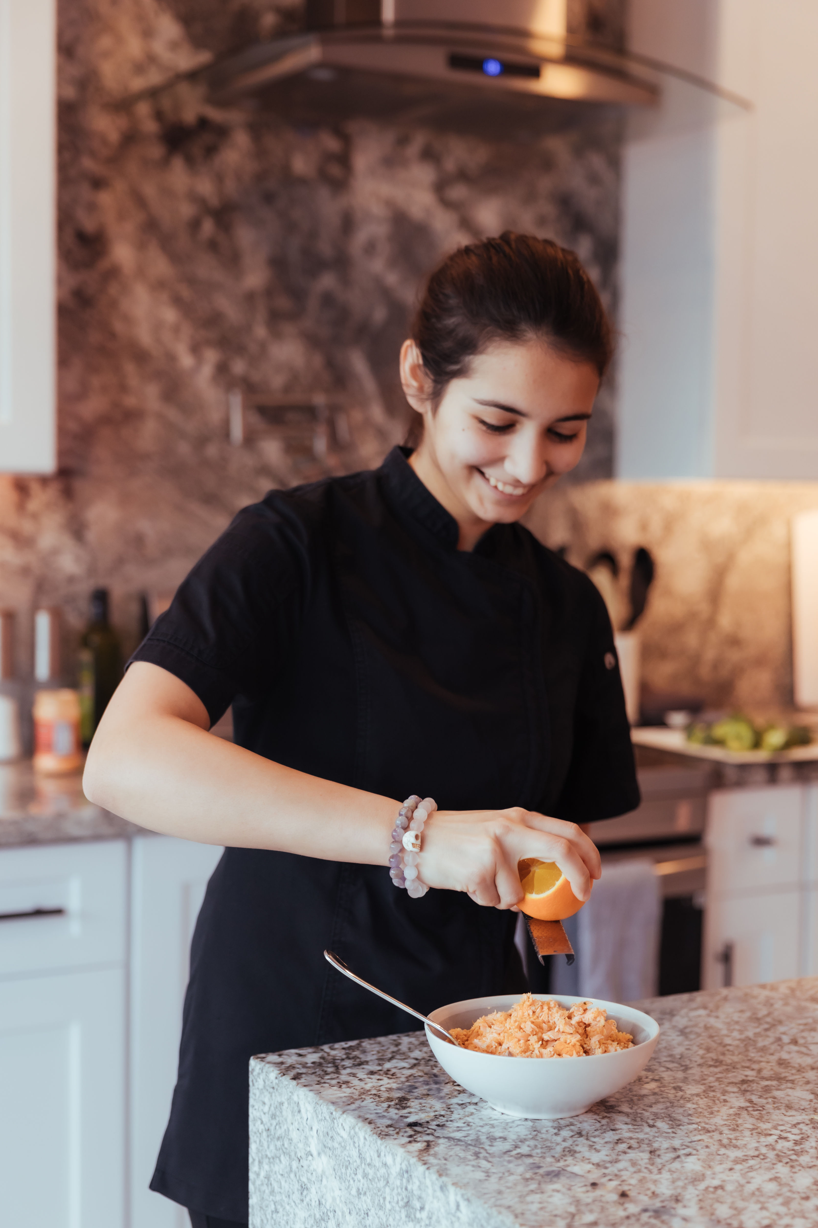 Smiling woman in a black chef jacket zesting an orange over a bowl of food on a granite kitchen island in a modern home kitchen.
