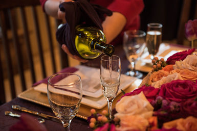 Close-up of champagne being poured into a flute at an elegant dining table with glassware and a pink-peach rose centerpiece — celebratory table setting