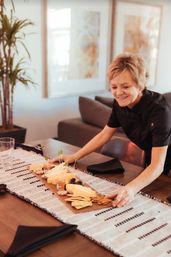 Smiling hostess arranging a wooden charcuterie board with cheeses, crackers and fruit on a modern dining table with a woven runner.