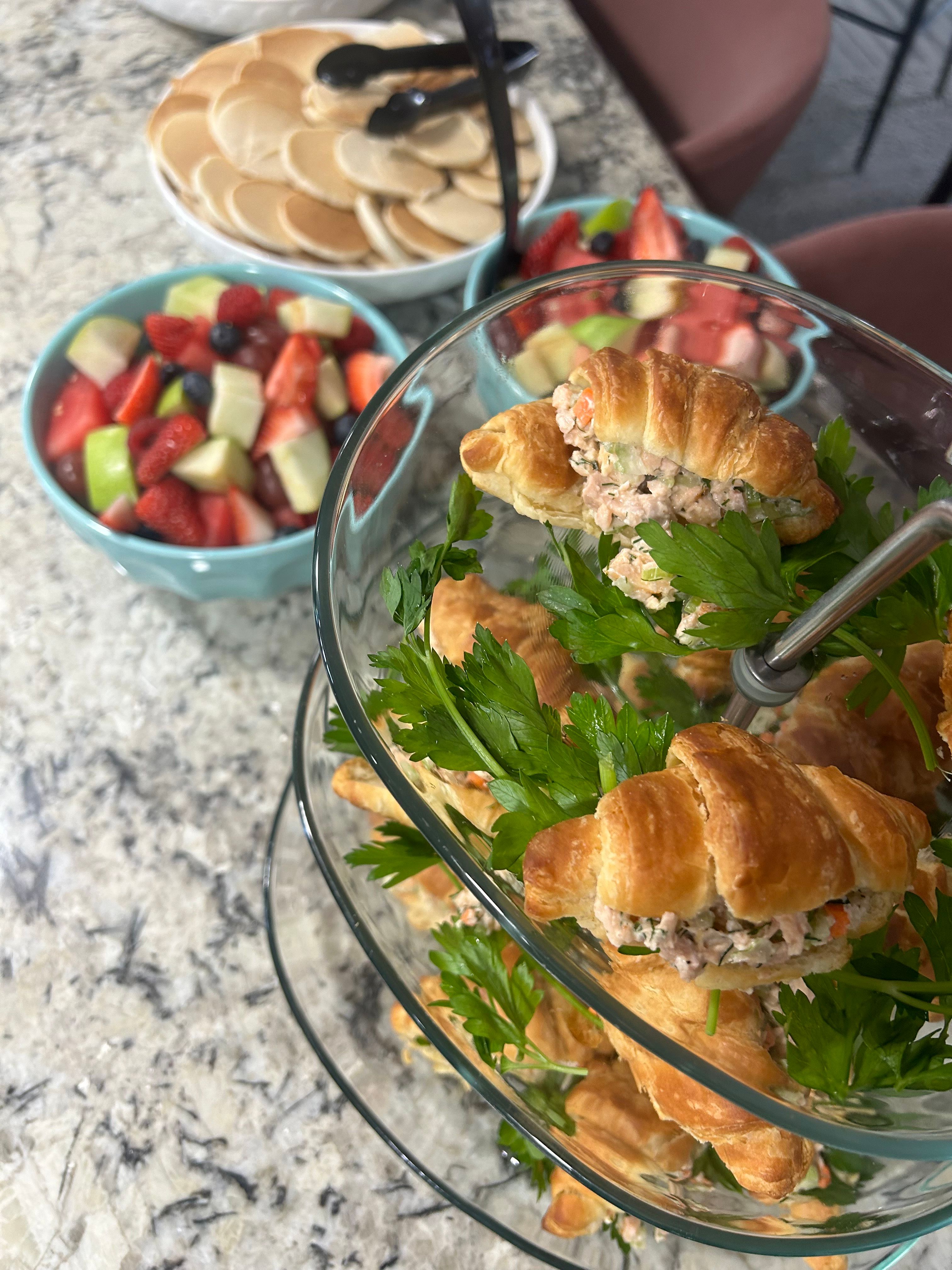 Tiered glass stand of mini croissant sandwiches filled with chicken salad and parsley, surrounded by bowls of mixed fruit (strawberries, melon, grapes, blueberries) and a plate of mini pancakes on a granite countertop — bright casual brunch spread.