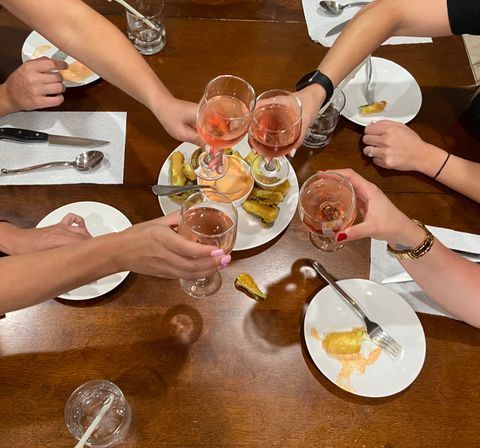Overhead shot of diners clinking rosé wine glasses in a cheers over a wooden table with a shared plate of fried appetizers and dipping sauce, small plates and utensils around.