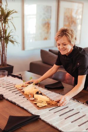 Smiling hostess arranging a wooden charcuterie board with cheeses, crackers and fruit on a modern dining table with a woven runner.