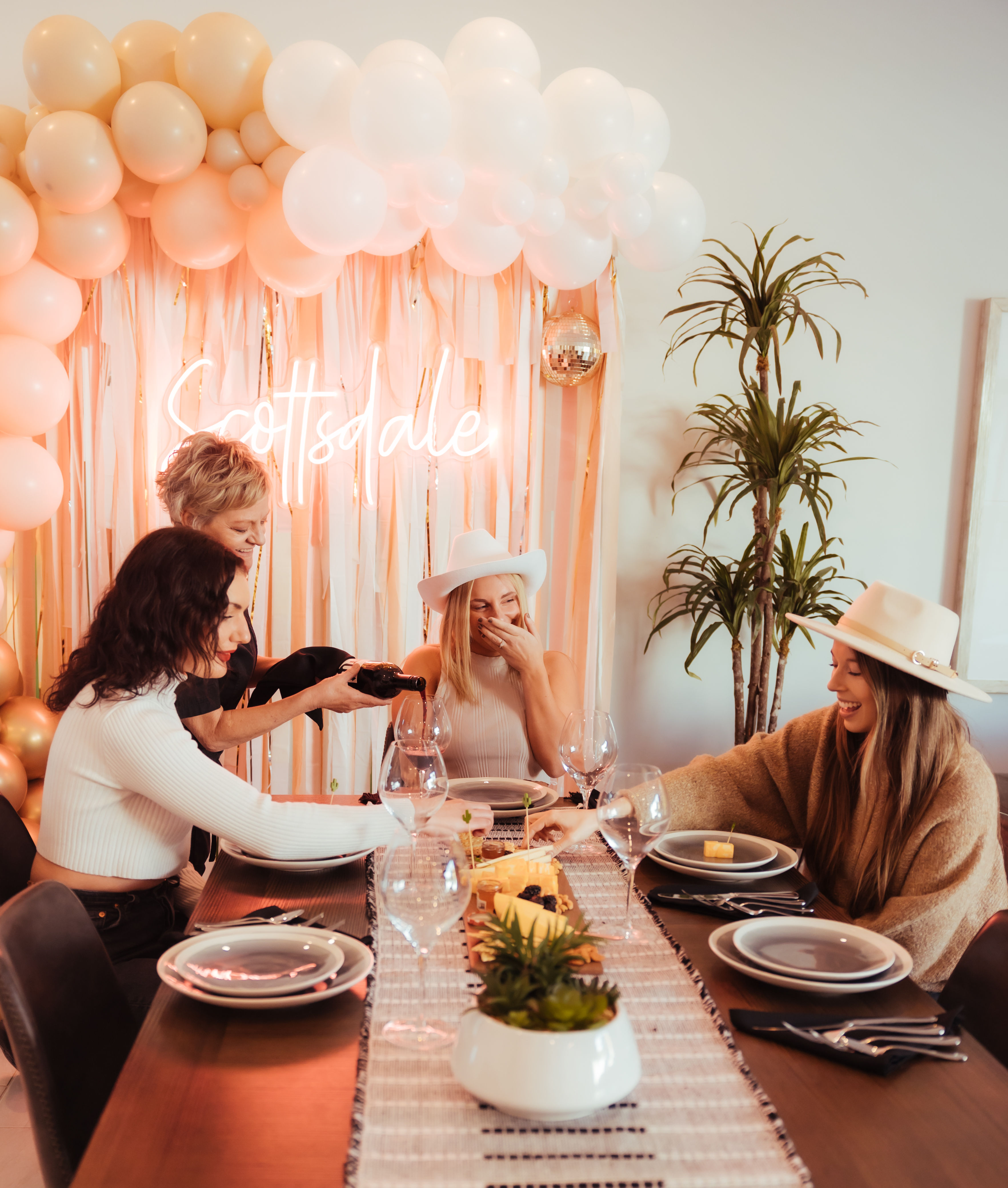 Four friends laughing at a festive indoor brunch with a pink-and-white balloon arch and neon 'Scottsdale' sign while wine is poured and a charcuterie board sits on the table.