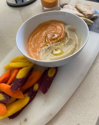 Swirled red-pepper and classic hummus drizzled with olive oil in a white bowl, served with purple, yellow and orange heirloom carrot sticks and pita on a marble board — vibrant Mediterranean snack on a kitchen counter.