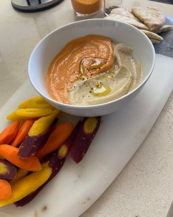 Swirled red-pepper and classic hummus drizzled with olive oil in a white bowl, served with purple, yellow and orange heirloom carrot sticks and pita on a marble board — vibrant Mediterranean snack on a kitchen counter.