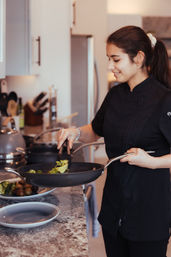 Smiling cook in a black chef jacket sautéing broccoli and mushrooms in a skillet on a modern kitchen countertop.
