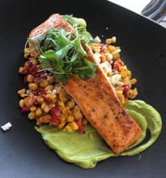 Seared salmon fillet on a bed of corn and red pepper succotash with a swipe of creamy avocado purée, crumbled white cheese and microgreens on a black plate.