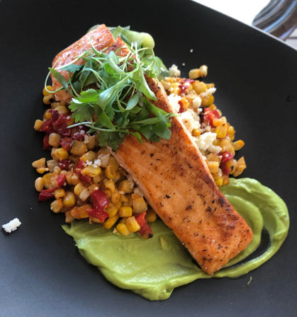 Seared salmon fillet on a bed of corn and red pepper succotash with a swipe of creamy avocado purée, crumbled white cheese and microgreens on a black plate.