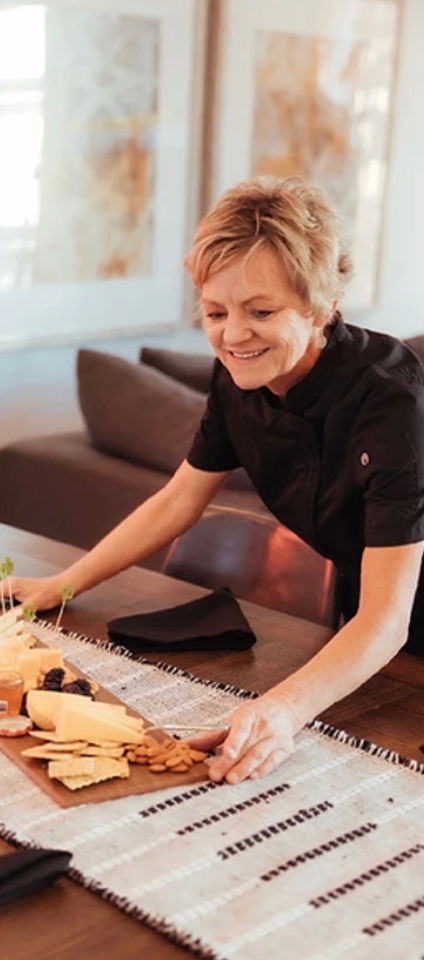 Smiling woman placing a cheese-and-cracker charcuterie board with nuts and spreads on a cozy dining room table