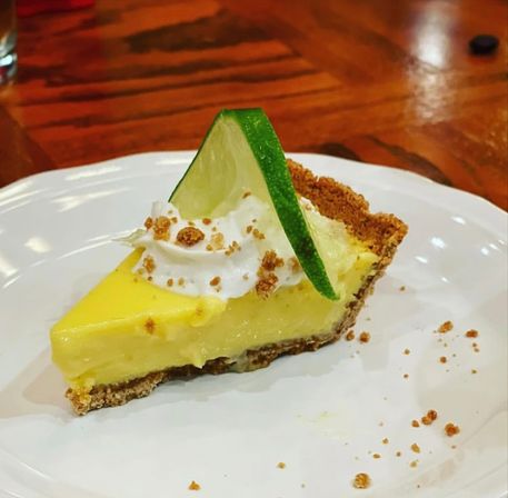 Zesty key lime pie slice with graham cracker crust, creamy yellow filling, whipped cream, lime wedge garnish and crumbs on a white plate on a wooden table