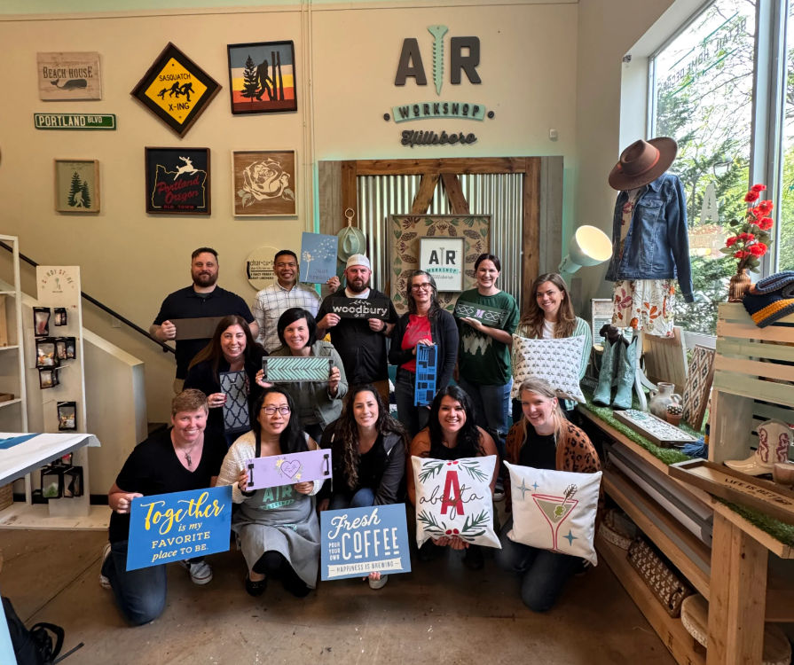 Thirteen people pose inside a bright Portland craft workshop, each holding handmade wood signs and decorative pillows amid rustic, Pacific Northwest-style decor and a large storefront window.