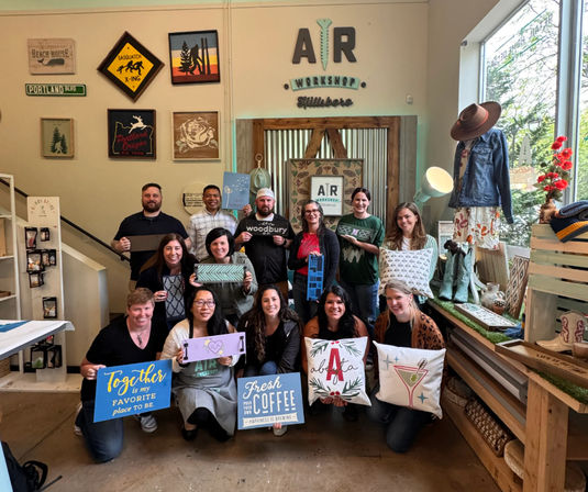 Thirteen people pose inside a bright Portland craft workshop, each holding handmade wood signs and decorative pillows amid rustic, Pacific Northwest-style decor and a large storefront window.