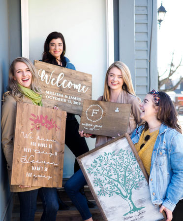 Four smiling friends on a porch outside a craft shop holding rustic wooden wedding signs and a painted family tree print, showcasing handmade wedding decor.
