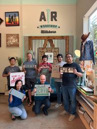 Group of seven people in a bright DIY craft studio posing with handmade wooden signs in front of a rustic corrugated metal backdrop, surrounded by framed art, a mannequin in a denim jacket, and shelves of home décor.
