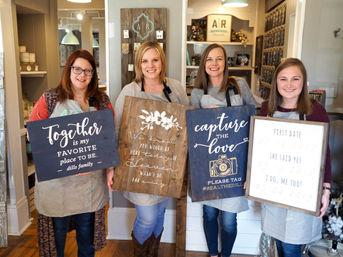 Four smiling women in aprons holding handmade wooden signs in a bright DIY sign-making workshop — rustic farmhouse-style signs with slogans and a camera graphic, shelving and craft supplies in the background.