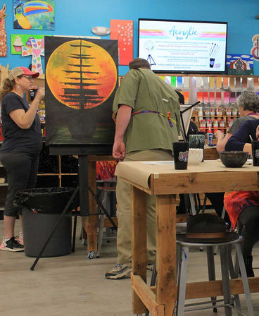 Acrylic painting class in a colorful art studio with participants working at wooden tables on a large canvas of a silhouetted tall ship against a glowing orange sunset, easel and paint supplies in the background.