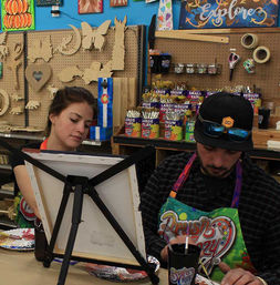 Two adults in a lively art studio workshop painting on canvases at an easel, colorful aprons and palettes on the table, pegboard wall with wooden cutouts and jars of paintbrushes in the background.