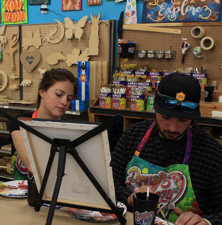 Two adults in a lively art studio workshop painting on canvases at an easel, colorful aprons and palettes on the table, pegboard wall with wooden cutouts and jars of paintbrushes in the background.
