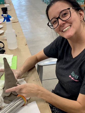 Smiling woman with glasses shaping a tall clay cone at a pottery class table in a ceramics studio, with a rolling pin and spray bottle nearby