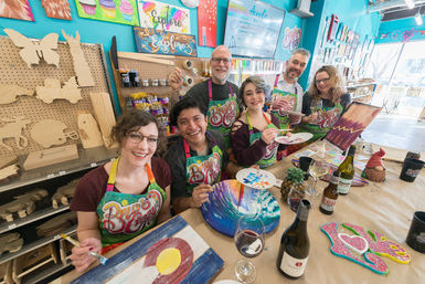 Six adults smiling at a paint-and-sip craft studio, wearing colorful aprons and painting wooden canvases including a Colorado-flag design and abstract pieces, with wine bottles, glasses, craft supplies and wooden cutouts on pegboard against bright turquoise walls.