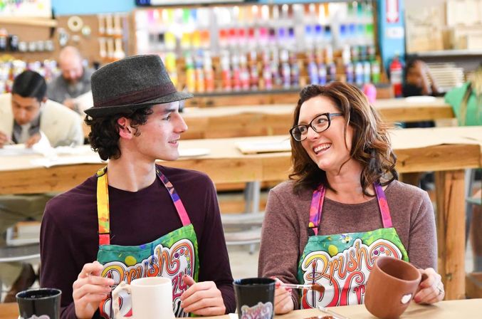 Two people in colorful aprons laughing as they paint ceramic mugs at a paint-your-own pottery studio, with shelves of paints and wooden tables in the background.