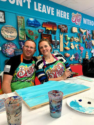 Smiling couple painting a turquoise wooden sign at a lively DIY craft-studio paint party, with paint cups and a palette on the table and a pegboard wall of colorful wooden shapes and signs behind them.