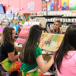 Four girls in a bright art studio wearing colorful aprons painting canvases — one canvas reads “Explore” over a watercolor world map, another shows a pink sunset, with shelves of blank ceramics and paint supplies in the background.
