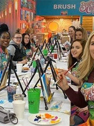 Paint-night group in an art studio: adults seated at a long table with tabletop easels, paintbrushes, palettes and drinks