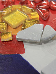 Close-up of colorful mosaic art detail: sparkly yellow glass tiles, red glass teardrops and silver ceramic shards on a red, white and blue painted background.
