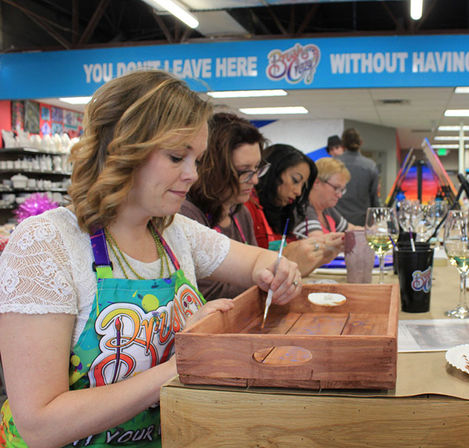 Group of women in colorful aprons at a paint-and-sip workshop painting wooden trays in a bright craft studio with wine glasses on the table