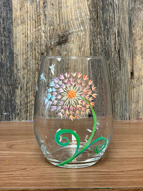 Hand-painted stemless wine glass featuring a colorful dandelion-style flower with fluttering petals and a green swirling stem, photographed on a wooden table with a rustic wood backdrop