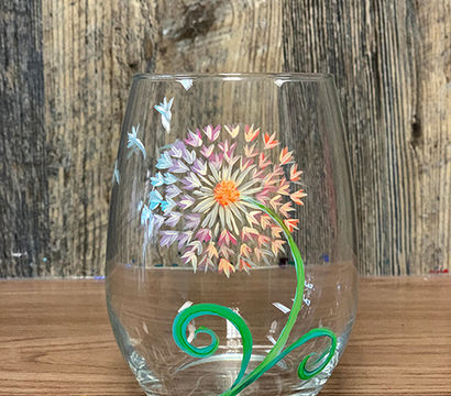 Hand-painted stemless wine glass featuring a colorful dandelion-style flower with fluttering petals and a green swirling stem, photographed on a wooden table with a rustic wood backdrop