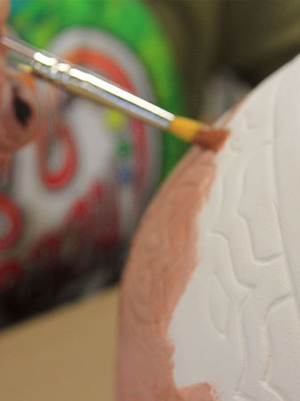 Close-up of a paintbrush applying peach paint to a white textured ceramic egg in a DIY craft project with a colorful blurred background