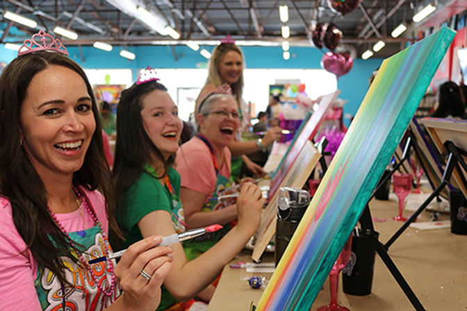 Smiling women at a lively indoor paint party in an art studio, wearing tiaras and aprons while painting colorful rainbow canvases on easels